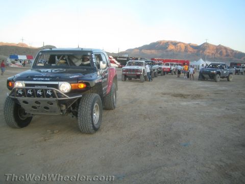 1990 Ford Bronco - black Toyota off-road race truck with a multi-light bumper leads a line of race trucks across a desert dirt lot.