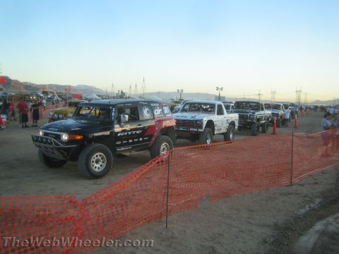 1990 Ford Bronco - line of off-road race trucks staged behind orange plastic safety fencing on dirt at dusk in the desert.