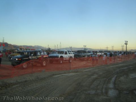 1990 Ford Bronco - long line of off-road race trucks staged behind orange safety fencing on a muddy dirt lot at dusk.