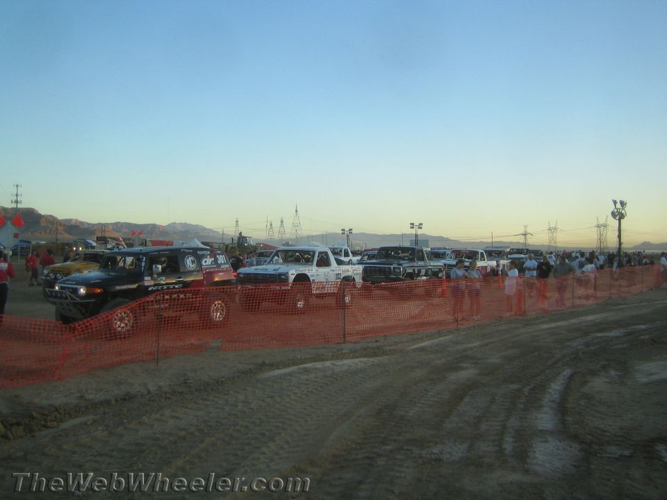 Long line of off-road race trucks behind orange fencing at dusk