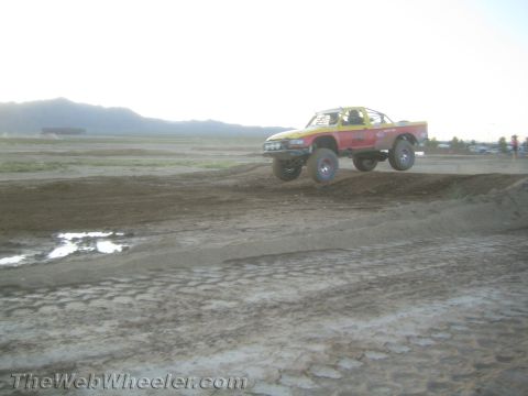 1990 Ford Bronco - red and yellow off-road race truck launches off a dirt mound on a short course with mountains in the background.