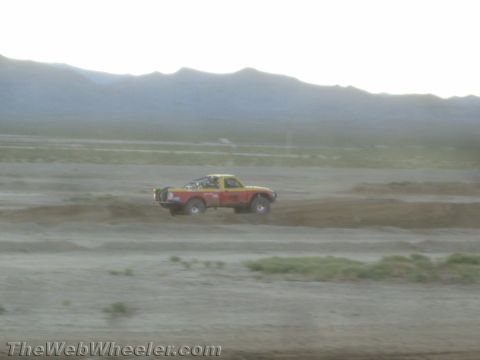 1990 Ford Bronco - red and yellow off-road race truck races across open desert terrain kicking up dust with mountains behind.