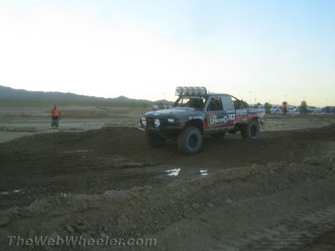 1990 Ford Bronco - black off-road race truck marked #743 sits on a graded dirt course as a track marshal stands nearby at dusk.