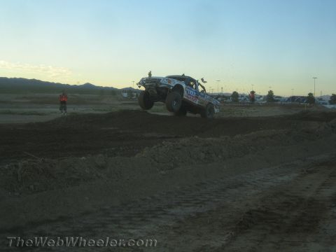 1990 Ford Bronco - off-road race truck launches off a dirt jump on a desert track in fading evening light.
