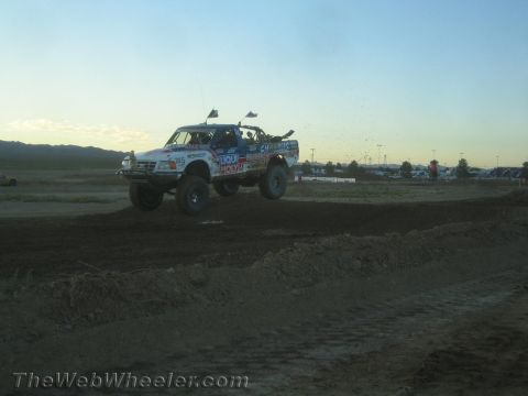 1990 Ford Bronco - white off-road race truck with sponsor decals flies over a dirt jump on a desert course at dusk.