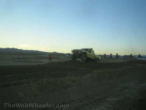 1990 Ford Bronco - bright yellow-green off-road race truck crests a dirt berm on a desert track as a marshal watches.