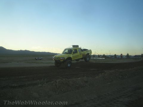 1990 Ford Bronco - yellow lifted off-road race truck rides the graded dirt track with mountains in the hazy background.