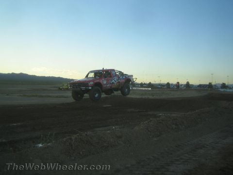 1990 Ford Bronco - red off-road race truck covered in decals catches air over a dirt jump on a desert course at dusk.