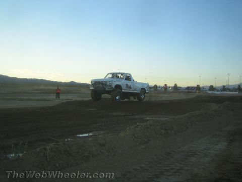 1990 Ford Bronco - white lifted pickup truck leaps over a dirt berm on a desert track in the evening.