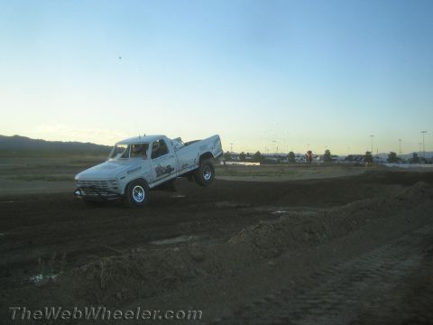 1990 Ford Bronco - white older-style pickup truck climbs a dirt jump on a graded desert course at dusk.
