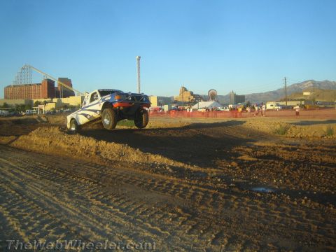 1990 Ford Bronco - white off-road race buggy crests a dirt berm with spectators and a building skyline behind it.