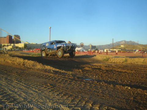 1990 Ford Bronco - blue Hummer H2 climbs a dirt berm on a desert off-road course with crowds and buildings behind.