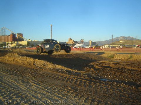 1990 Ford Bronco - off-road race truck flies over a dirt jump on a desert course with spectators lining the track.