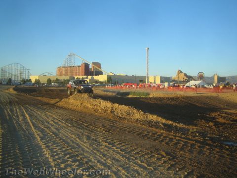 1990 Ford Bronco - red and black truck crosses a dirt berm on a desert course with a roller coaster and crowds behind.