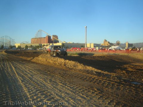 1990 Ford Bronco - dark pickup truck rides a dirt berm on a graded desert course with a fairground and roller coaster behind.
