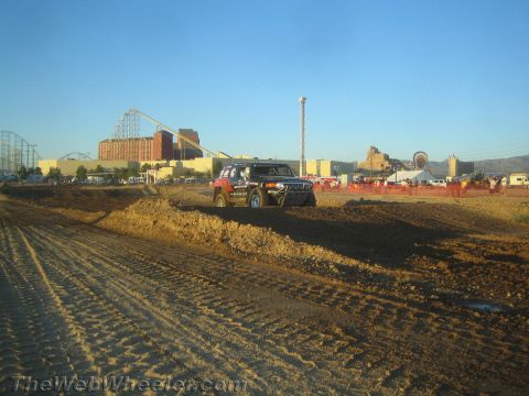 1990 Ford Bronco - boxy decaled off-road SUV navigates a dirt course with buildings and a roller coaster in the background.