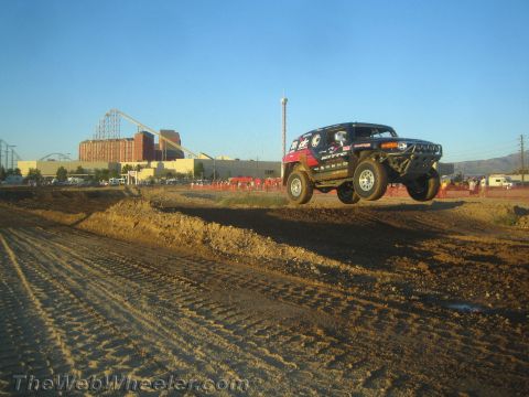 1990 Ford Bronco - boxy off-road SUV catches air over a dirt jump on a desert course with a roller coaster behind.