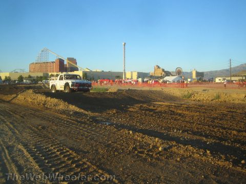 1990 Ford Bronco - white pickup truck climbs a dirt berm on a graded desert course with crowds and a roller coaster behind.