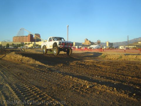 1990 Ford Bronco - white off-road race truck flies over a dirt jump on a desert course with a roller coaster in the background.
