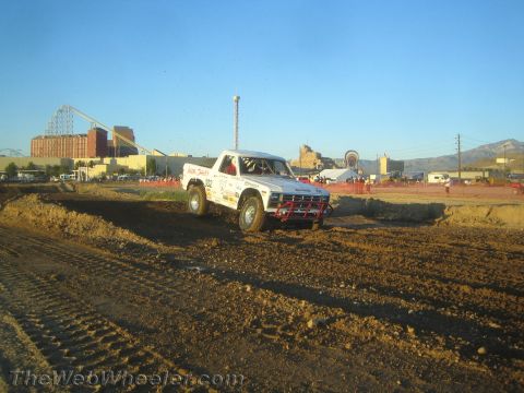 1990 Ford Bronco - white short-course off-road race truck digs through a banked dirt corner with an amusement park roller coaster on the horizon.