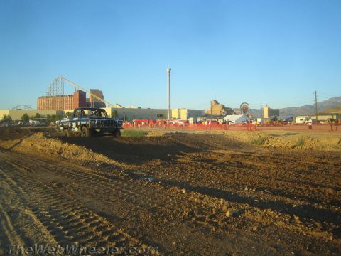 1990 Ford Bronco - dark pickup truck sits on a churned dirt off-road course with a roller coaster and industrial buildings in the background.