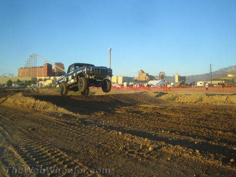 1990 Ford Bronco - black off-road race truck catches air over a dirt jump with a roller coaster and mountains in the background at golden hour.