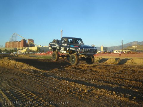 1990 Ford Bronco - black off-road race truck numbered 300 jumps a dirt berm with roller coaster and mountains behind in evening light.