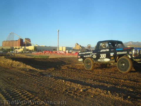 1990 Ford Bronco - black off-road race truck marked 300 with West Coast Broncos and BFGoodrich decals corners on a dirt track at golden hour.