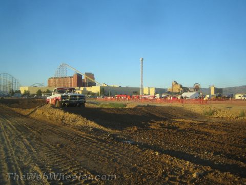 1990 Ford Bronco - graded dirt off-road course with a light-colored truck staged at left and a roller coaster and crowd in the distance.