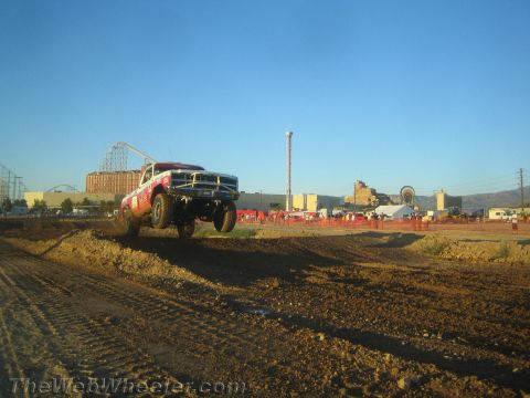 1990 Ford Bronco - red and white off-road race truck flies over a dirt jump with a roller coaster and mountains behind in evening light.