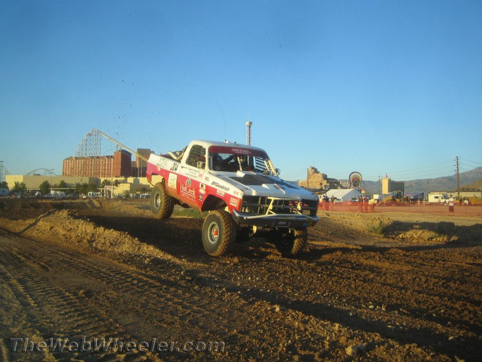 Red and white race truck landing a dirt jump with checkered hood at dusk