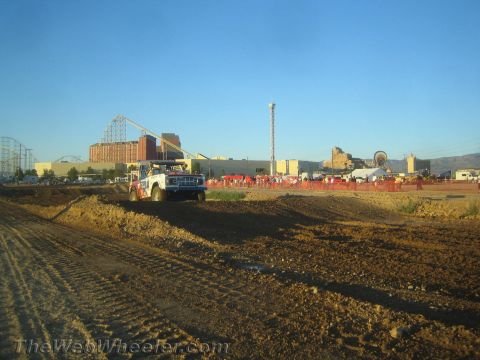 1990 Ford Bronco - red and white off-road race truck works along a dirt course with a roller coaster, buildings and a crowd in the distance.
