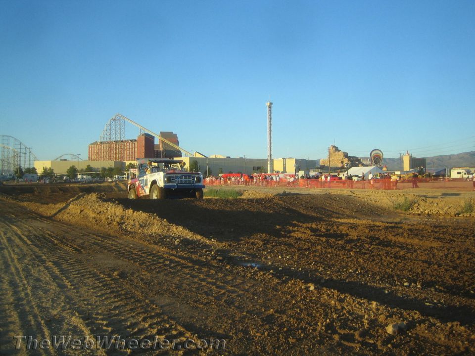 Red and white off-road race truck on a dirt course, roller coaster behind