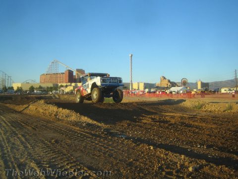 1990 Ford Bronco - blue and white off-road race truck launches off a dirt berm with a roller coaster and mountains behind in evening light.