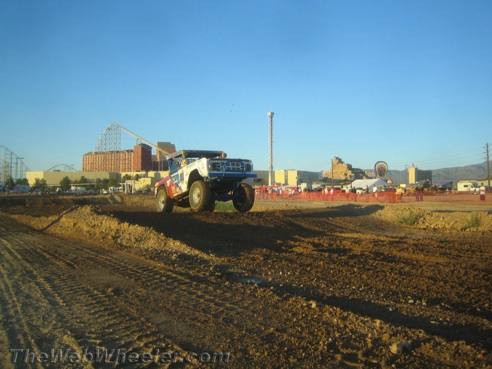 Blue and white off-road race truck airborne over a dirt berm at dusk