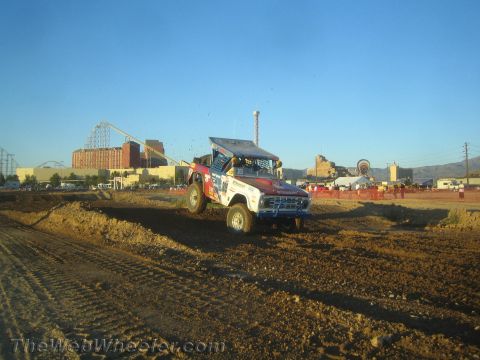 1990 Ford Bronco - blue and white off-road race truck crests a dirt rise on the track with a roller coaster and buildings in the background.