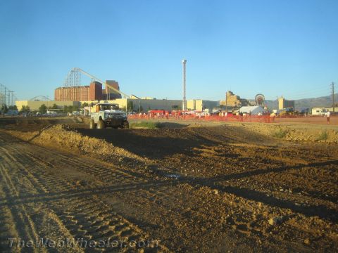 1990 Ford Bronco - light-colored off-road truck moves along a graded dirt course with a roller coaster, buildings and a crowd in the distance.