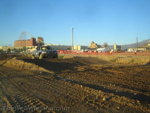 1990 Ford Bronco - lifted off-road pickup truck climbs a dirt track berm with roller coasters and a fairground visible behind it at dusk.