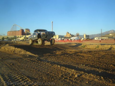 1990 Ford Bronco - lifted off-road pickup truck catches air over a dirt berm with an amusement park and mountains in the background.