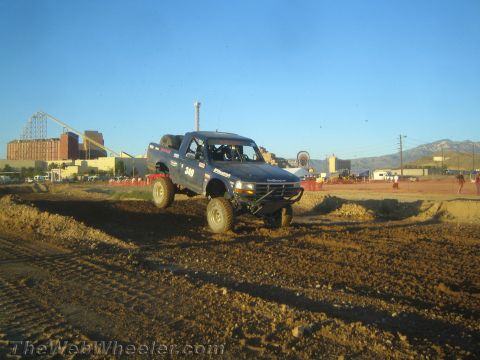 1990 Ford Bronco - off-road race truck marked 349 climbs over a dirt berm with a fairground and mountains behind it.