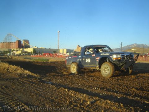 1990 Ford Bronco - blue off-road pickup truck numbered 349 drives over a dirt berm with mountains and a fairground in the distance.
