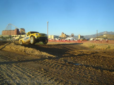 1990 Ford Bronco - yellow off-road pickup truck drives over a dirt berm while spectators line the course near a fairground.