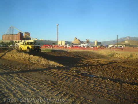 1990 Ford Bronco - yellow off-road pickup truck parked on a churned dirt track with industrial buildings and an amusement park behind it.
