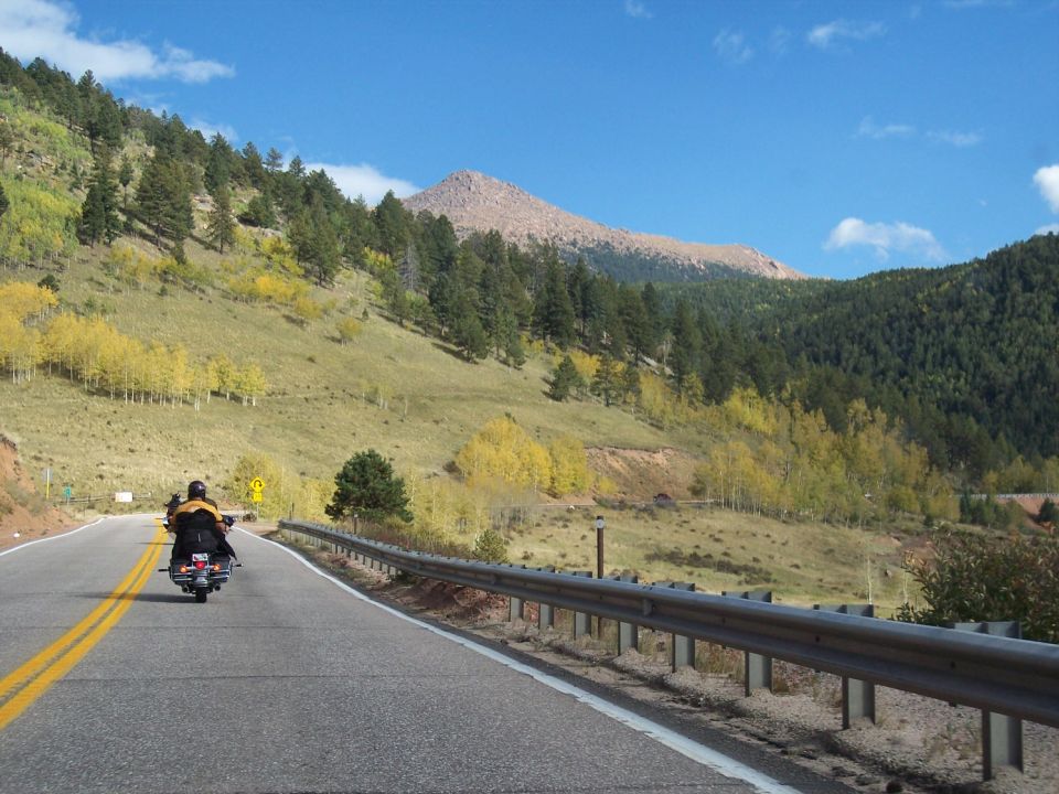 Motorcycle riding on a paved road with yellow double lines, surrounded by hills with autumn foliage and pine trees under a blue sky