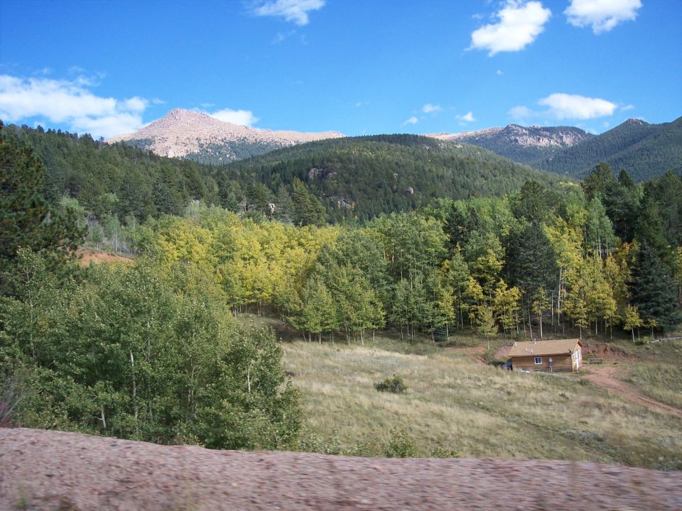 Cabin on a grassy slope with trees and mountains under a blue sky