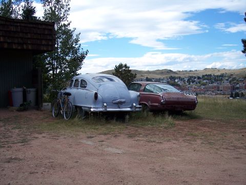 1972 Ford Ranchero - Two vintage cars sit parked in a grassy area next to a building, with a town visible on a distant hillside.