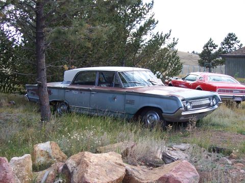 1972 Ford Ranchero - A weathered 1963 Mercury Monterey sedan sits in a grassy area next to a red car, showing its distinctive grille and two-tone paint.
