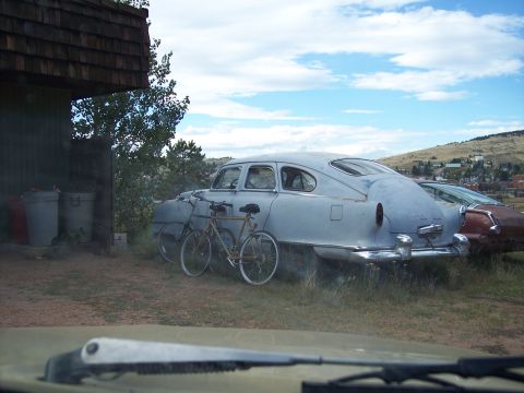 1972 Ford Ranchero - Two bicycles lean against a light blue sedan parked in a grassy area next to a building.
