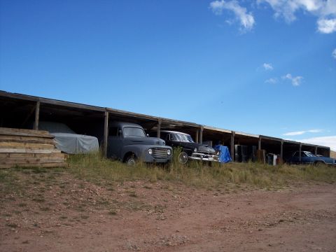 1972 Ford Ranchero - Several old vehicles are parked under a long wooden shelter on a grassy lot under a blue sky.