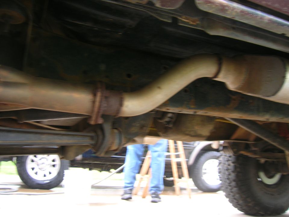 Underside of a vehicle showing exhaust pipes and suspension components, with a person in blue jeans visible